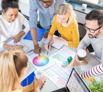 Team collaborating around a table, pointing to a color wheel and analytics charts during a design planning session.