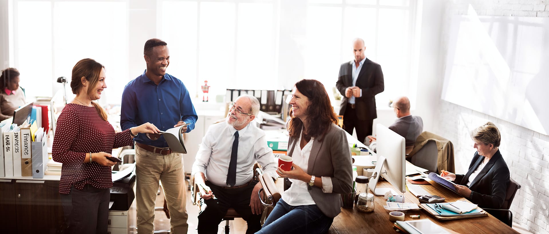 Priority-Staffing-Business-Casual Coworkers in a bright open office chat and review papers around a long desk, while one person sits holding a red coffee cup.