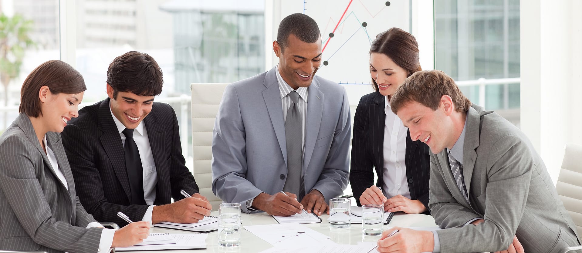 Priority-Staffing-Business-Professional Five colleagues in suits sit at a conference table, smiling and taking notes, with a chart on a board behind them.