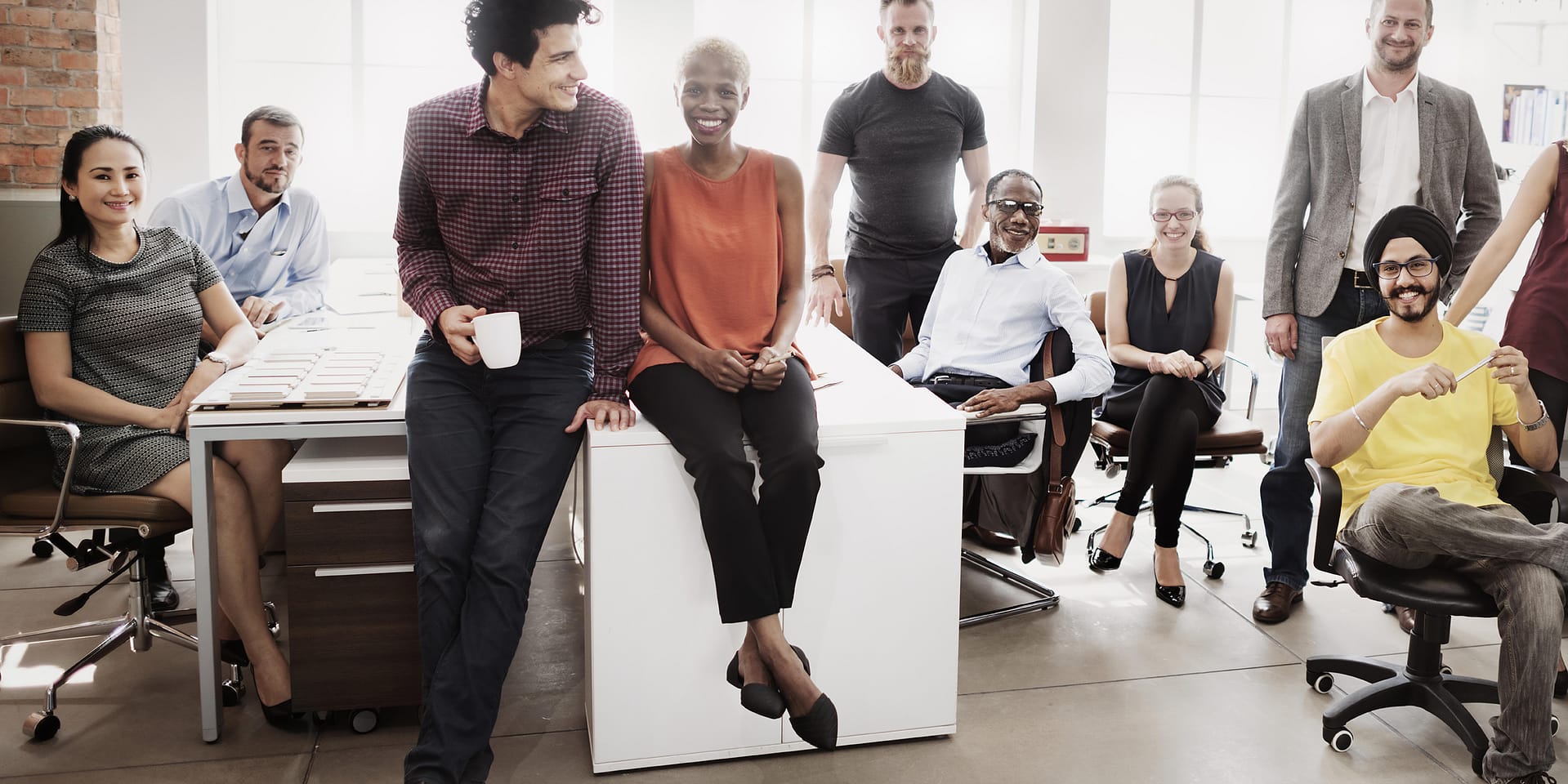 Priority-Staffing-Casual-Workwear Group of coworkers pose together in a bright office, smiling, with some seated and others standing around desks and chairs.