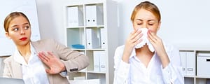 One person sneezes into a tissue at an office desk while a nearby coworker leans back with a hand raised to avoid it.
