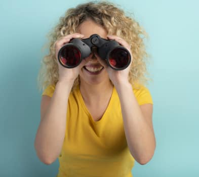Job seekers Curly-haired person in a yellow T-shirt smiles while looking through black binoculars against a light blue background.