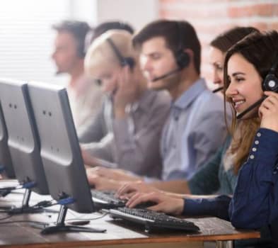 Two women working at desks in a call center, engaged in conversation and using headsets.
