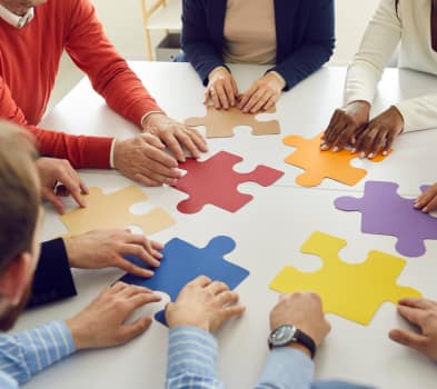 Team members arrange large, colorful puzzle pieces on a table, many hands reaching in to assemble a solution.