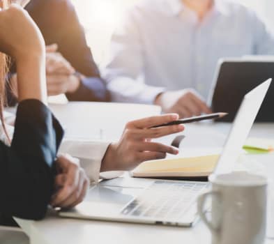 Close-up of colleagues at a bright meeting table; a hand with a pen points to a laptop beside a notebook and coffee mug.