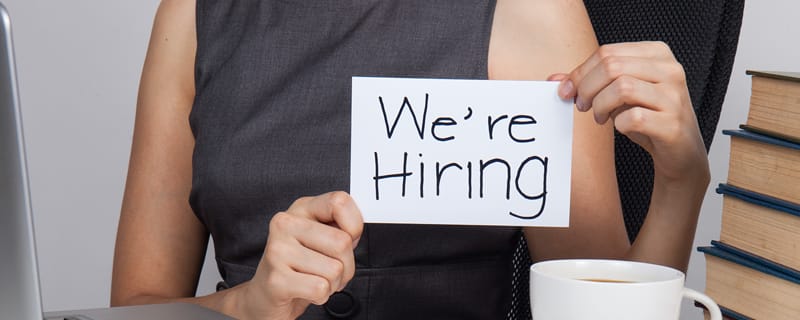Person holds a handwritten sign reading “We’re Hiring” at a desk with a coffee cup and stacked books.