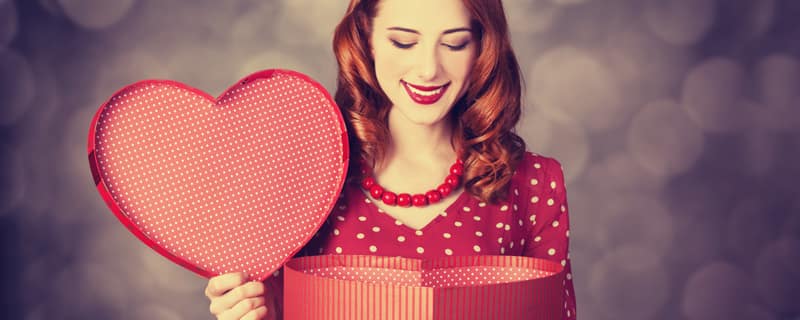 Woman in a red polka-dot dress smiles while opening a red gift box, holding a heart-shaped lid against a soft bokeh backdrop.