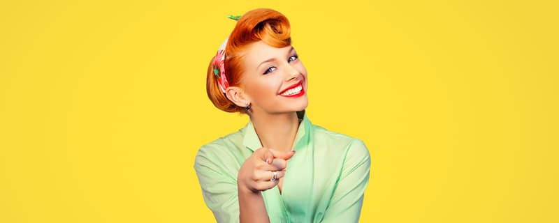 Smiling person with a retro hairstyle points toward the camera, wearing a mint blouse against a bright yellow background.