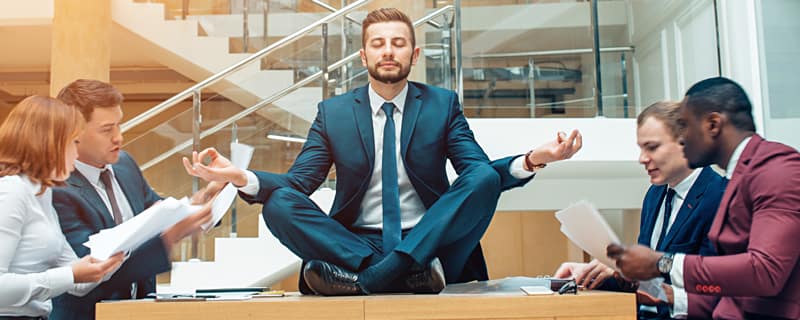 Person in a suit sits cross-legged meditating on a desk as coworkers around shuffle papers in a busy office atrium.